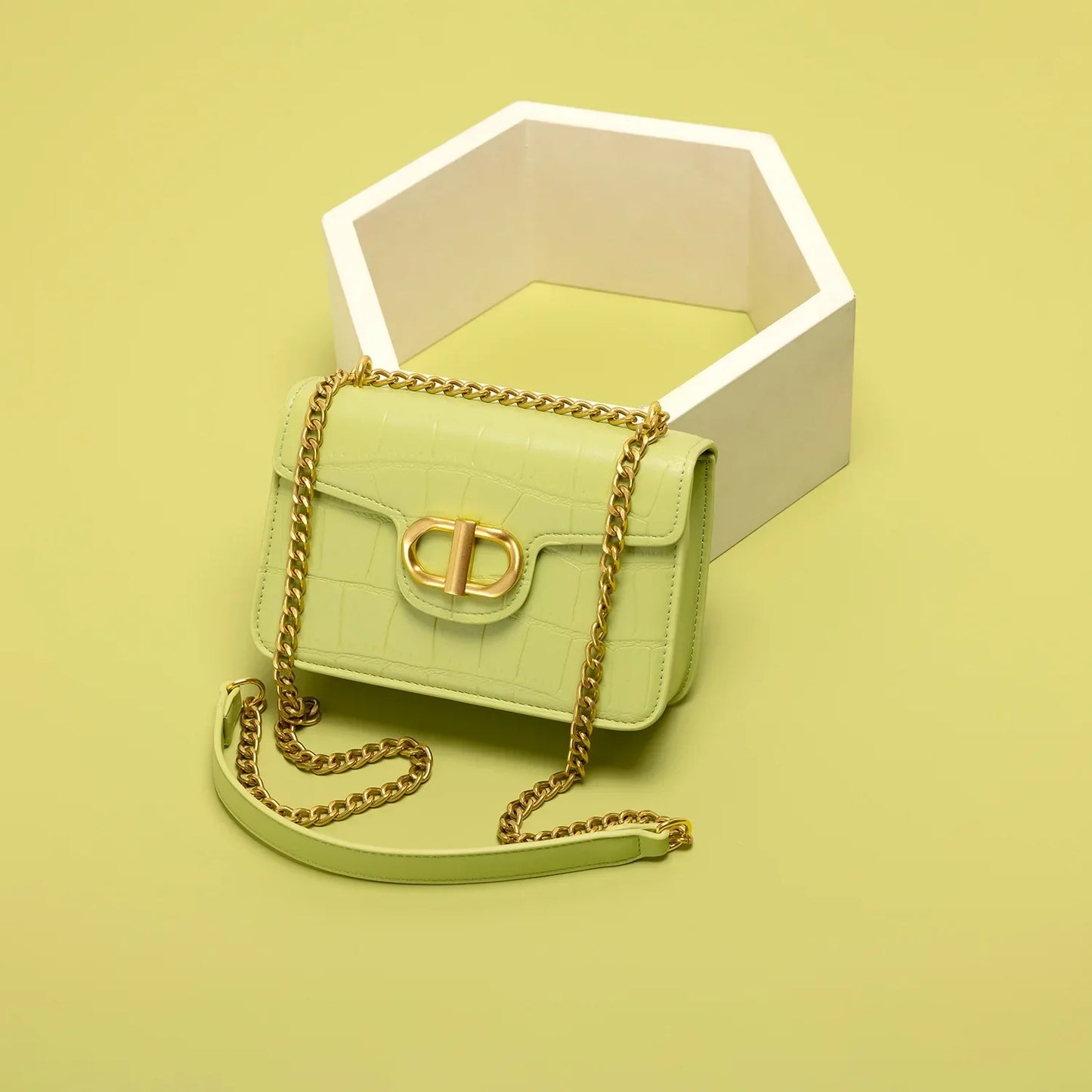 Light green handbag with gold chain strap, placed next to a white hexagonal decoration on a soft yellow background.